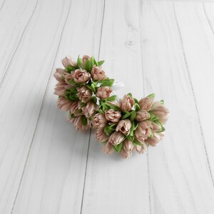 May include: Two bouquets of artificial tulips in a soft peach color with green leaves. The flowers are clustered together, creating a full and textured appearance. The background is a light, wooden surface.