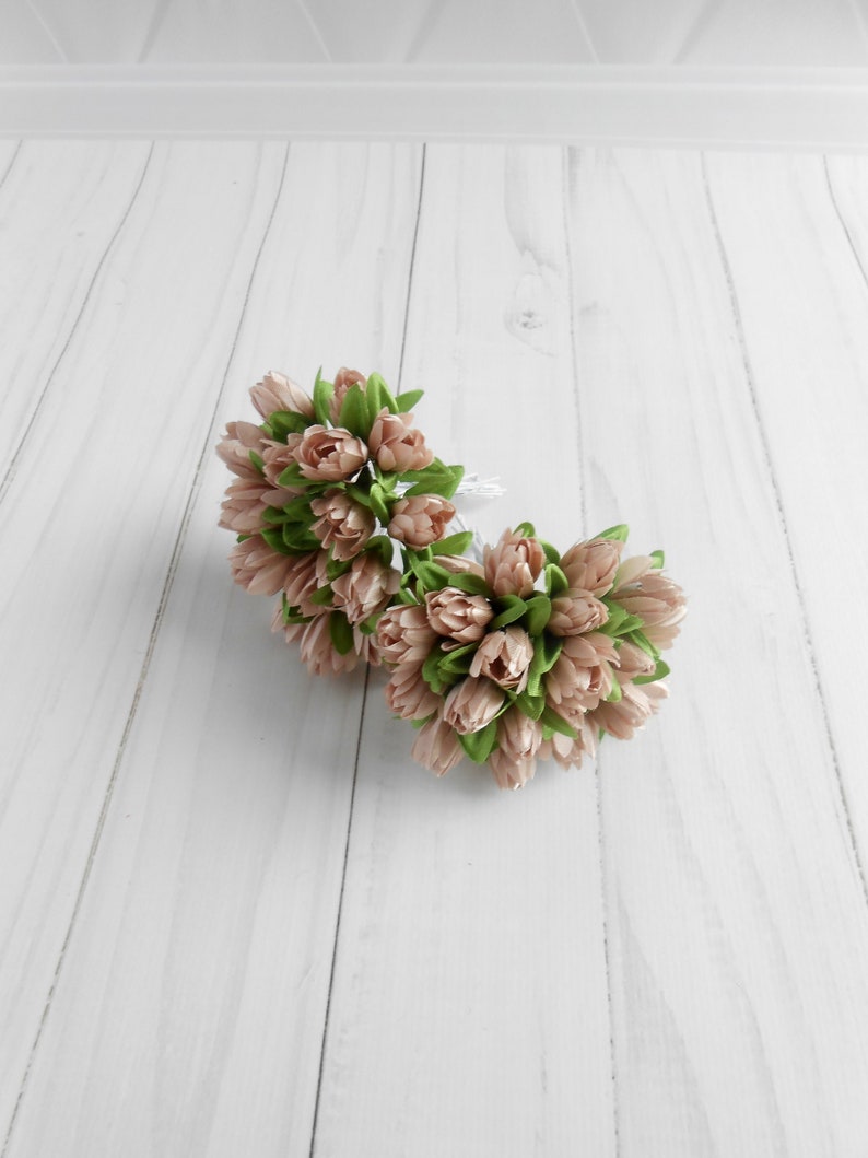 May include: Two bouquets of artificial tulips in a soft peach color with green leaves. The flowers are clustered together, creating a full and textured appearance. The background is a light, wooden surface.
