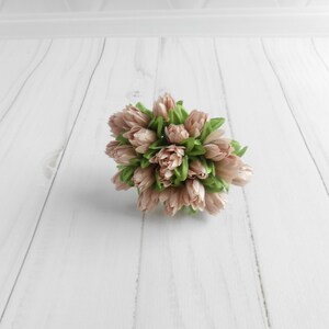 May include: A cluster of artificial tulips in shades of blush pink and green. The faux flowers are arranged in a compact, rounded shape, with the pink petals tightly closed. The green leaves peek out from the base of the blooms. The arrangement is set against a white background.