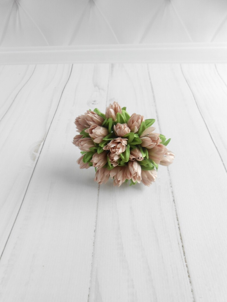 May include: A cluster of artificial tulips in shades of blush pink and green. The faux flowers are arranged in a compact, rounded shape, with the pink petals tightly closed. The green leaves peek out from the base of the blooms. The arrangement is set against a white background.