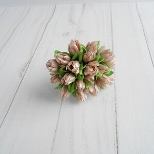 May include: A cluster of artificial flowers with light pink petals and green leaves. The arrangement is tightly packed, creating a rounded shape. The flowers are set against a white wooden background.
