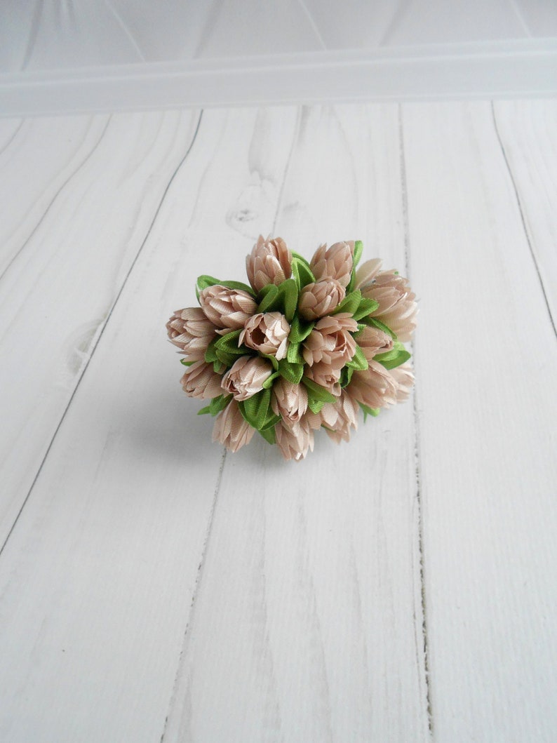 May include: A cluster of artificial flowers with light pink petals and green leaves. The arrangement is tightly packed, creating a rounded shape. The flowers are set against a white wooden background.