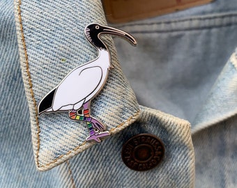 May include: A white and black bird pin with a long, curved beak and rainbow-coloured legs is attached to a light blue denim jacket. The pin is a stylized representation of an ibis.