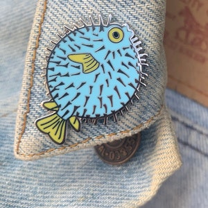 May include: A blue and yellow enamel pin of a pufferfish with spikes. The pin is on a denim jacket.