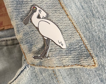 May include: A silver enamel pin depicting a white bird with a long, black beak. The pin is displayed on a denim background.
