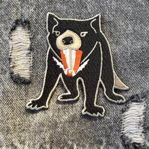 May include: An embroidered patch of a Tasmanian devil, in black and white, with a large, open mouth showing sharp teeth. The patch is sewn onto a piece of denim fabric.