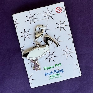May include: A silver zipper pull featuring a white ibis bird with black accents. The zipper pull is on a white card with a silver star pattern and the text "Zipper Pull Bush Bling Australia bushbling.com.au".