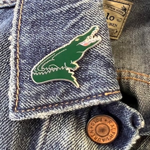 May include: A green alligator enamel pin on a blue denim jacket. The pin is a detailed depiction of an alligator with a toothy grin.
