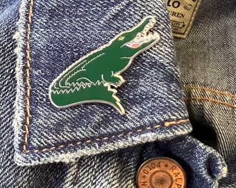 Puede incluir: Un pin de esmalte de cocodrilo verde en una chaqueta vaquera azul. El pin es una representación detallada de un cocodrilo con una sonrisa de dientes.
