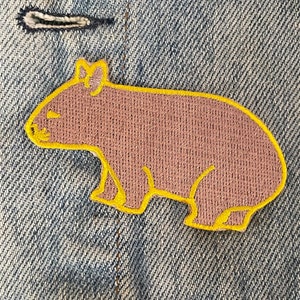 Könnte beinhalten: Ein brauner gestickter Aufnäher mit einem Cartoon-Wombat mit gelber Umrandung auf einem blauen Jeansstoff.