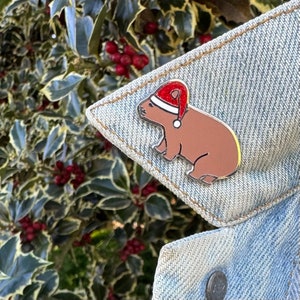 May include: A brown capybara wearing a red Santa hat with white trim. The capybara is sitting on a blue denim jacket.
