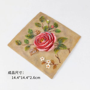 May include: A square, beige-colored tile with a 3D sculpted design of a pink rose with green leaves and white flowers. The tile measures 14.4 x 14.4 x 2.6 inches.
