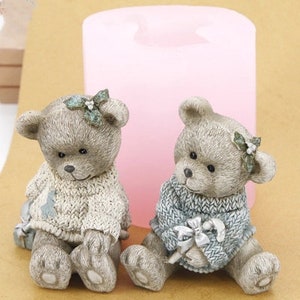 May include: Two ceramic teddy bear figurines wearing knitted sweaters. The bear on the left is wearing a cream sweater with a blue and white design. The bear on the right is wearing a blue sweater with a silver bow.
