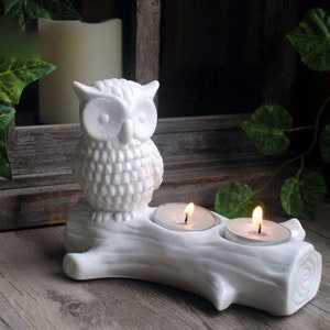 May include: White ceramic owl candle holder with two candle wells. The owl is perched on a tree branch with a rustic wood grain texture.