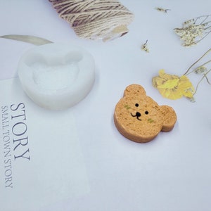 May include: A white silicone mold shaped like a bear's head. A brown bear-shaped cookie is next to the mold.
