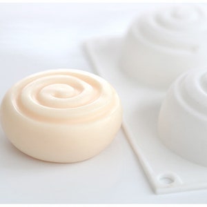 K&ouml;nnte beinhalten: Ein hellbeige, spiralf&ouml;rmige Seife liegt vor zwei wei&szlig;en Seifenformen. Die Seife hat eine glatte, abgerundete Form mit einem Spiraldesign auf der Oberseite. Die Formen sind ebenfalls spiralf&ouml;rmig, was auf ein Seifenherstellungsthema hindeutet.