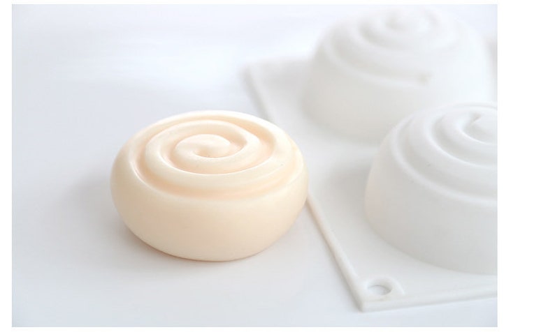 K&ouml;nnte beinhalten: Ein hellbeige, spiralf&ouml;rmige Seife liegt vor zwei wei&szlig;en Seifenformen. Die Seife hat eine glatte, abgerundete Form mit einem Spiraldesign auf der Oberseite. Die Formen sind ebenfalls spiralf&ouml;rmig, was auf ein Seifenherstellungsthema hindeutet.