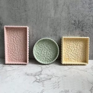 May include: Three decorative trays in different shapes and colors. A rectangular tray is light pink, a round tray is light green, and a square tray is light yellow. Each tray has a textured, pebble-like pattern.