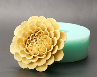 May include: A yellow silicone mold for making a flower-shaped cake decoration. The mold is sitting on a gray surface and a yellow flower-shaped cake decoration is sitting next to the mold.