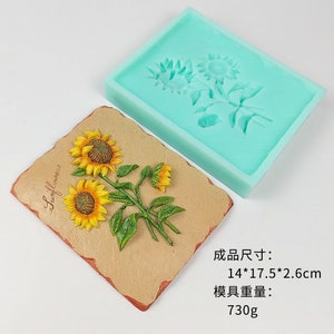 May include: A silicone mold for making a sunflower cake topper. The mold is light blue and shows three sunflowers. The finished product is shown next to the mold and is 14 by 17.5 by 2.6 inches and weighs 730 grams.