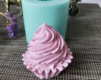 May include: A light purple cupcake made of soap with a detailed swirl design, next to a teal silicone mold. The cupcake sits on a gray surface. The mold is cylindrical with a ridged interior.