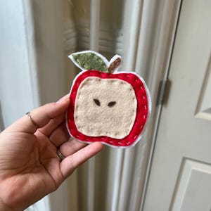 May include: A felt apple-shaped ornament with a red exterior, white stitching, and a tan interior. The apple has a green leaf and a brown stem. The ornament is held in a hand.