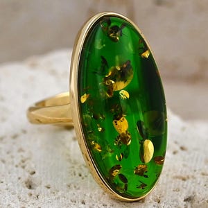 May include: A gold ring with a large, oval green gemstone with golden flecks. The ring has a polished gold band and bezel setting. The gemstone's colour is a vibrant, translucent green, creating a striking contrast with the gold.