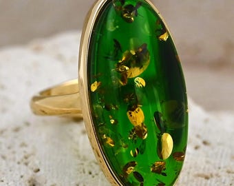 Pode incluir: Um anel de ouro com uma grande pedra verde oval com inclusões douradas. O anel tem um design simples e elegante com uma banda de ouro polido e uma moldura. A pedra tem uma cor verde vibrante e translúcida.