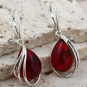 Peut inclure: Boucles d'oreilles pendantes argentées avec des pierres rouges en forme de goutte d'eau. Les pierres sont serties dans un design en fil d'argent. Les boucles d'oreilles ont une fermeture à levier. Elles sont présentées sur une surface texturée de couleur claire.