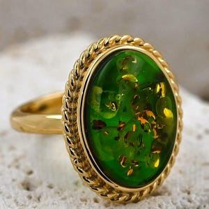 May include: A gold ring with an oval, green gemstone with golden flecks. The gemstone is framed by a twisted rope design. The ring's band is plain, polished gold.