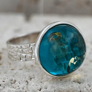 May include: A silver ring featuring a large, round, teal stone with gold inclusions. The stone is set in a silver bezel. The ring's band has a textured, brick-like design.