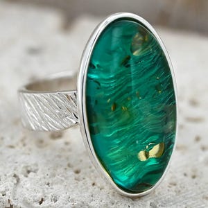 May include: A silver ring with a large, oval, translucent green stone. The stone has a swirling pattern with gold flecks. The ring band has a textured, etched design.