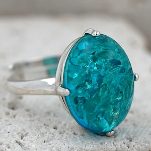 May include: A silver ring with a large, oval teal gemstone. The stone has a textured, crystalline look, set in a simple silver band. The ring is shown on a light, textured surface. The gemstone's dimensions are approximately 2.5 cm in length.