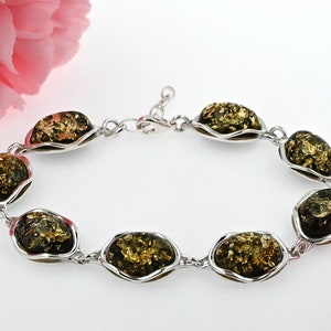Könnte beinhalten: Ein silbernes Armband mit grünen Bernstein-Steinen. Die Steine sind oval geformt und haben einen goldenen Schimmer. Das Armband hat einen Karabinerverschluss.