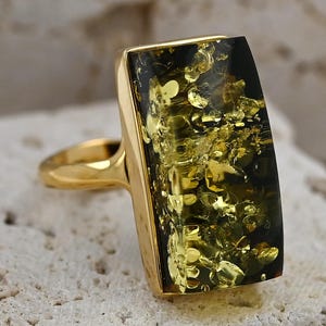 May include: A gold-toned ring featuring a rectangular, dark green amber stone with golden inclusions. The ring has a simple, elegant design with a smooth, polished finish. The amber stone is set in a gold bezel.