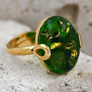 May include: A gold ring featuring a large, oval green stone with gold flecks. The ring has a polished gold band and setting. The design includes decorative loops on the sides. The stone's colour is a vibrant green, and the overall design is elegant.