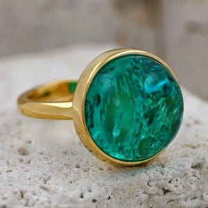 May include: A gold ring featuring a large, round, emerald-green stone. The stone has a textured, marbled appearance. The ring band and setting are a bright, polished gold colour. The ring is set on a textured, off-white surface.