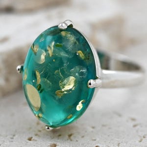 Peut inclure: Une bague en argent avec une grande pierre précieuse ovale de couleur verte sertie dans une bélière en argent. La pierre précieuse est parsemée de paillettes dorées.