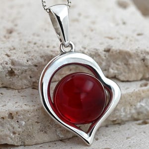 Può includere: Una collana con pendente in argento con una cornice a forma di cuore che contiene una gemma rotonda di colore rosso vivo. Il pendente è sospeso a una sottile catena d'argento. La gemma ha un diametro di circa 2 cm.