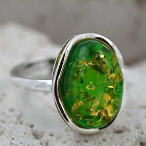 May include: Oval ring featuring a bright green stone with gold inclusions, set in a silver band. The ring is presented on a textured, light-coloured surface. A piece of jewellery.
