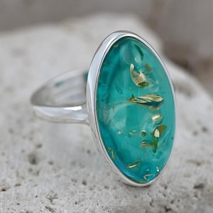 May include: A silver ring featuring a large, oval turquoise stone with gold inclusions. The stone is set in a silver band. The ring is presented on a textured, light-coloured surface.