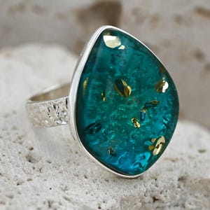 May include: A silver ring with a large, teardrop-shaped teal stone. The stone is embedded with gold flecks and set in a silver bezel. The ring band has a textured, hammered design.