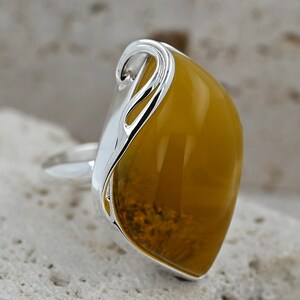 May include: A silver ring featuring a large, teardrop-shaped amber stone. The amber is a warm, golden-yellow color with natural inclusions. The ring has a polished silver band and a decorative silver setting that curves around the amber.