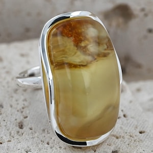 May include: A silver ring featuring a large, rectangular amber stone. The amber has a warm, golden-yellow hue with darker, swirling patterns. The ring's setting is a polished silver, creating a striking contrast with the amber.