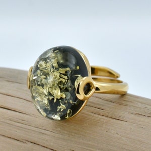 May include: A gold ring featuring a large, round, green amber stone set in a gold bezel. The stone has a black and green marbled pattern.