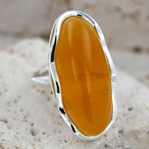 May include: A silver ring featuring a large, oval-shaped amber-colored stone. The stone is set in a polished silver band with a unique, wavy border. The ring is displayed on a textured, light-colored surface.