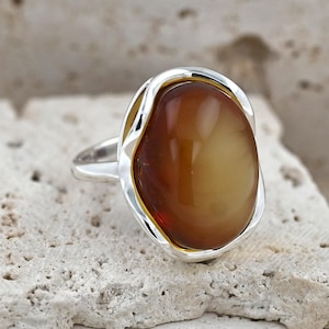 May include: A silver ring featuring a large, smooth, oval-shaped amber gemstone with a deep cognac color. The metal setting has a polished finish and wraps around the edges of the stone, creating a continuous, flowing frame around the amber.
