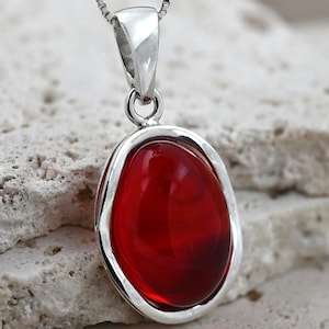 Puede incluir: Un collar con un colgante de plata con una piedra ovalada roja. El colgante tiene un borde liso y redondeado y un acabado pulido.