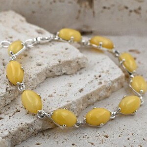 Op de afbeelding: Een zilveren armband met ovale gele barnsteenstenen. De armband ligt op een wit, getextureerd oppervlak.