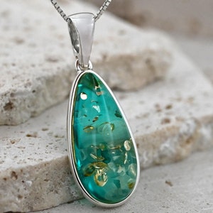 May include: A teardrop-shaped pendant with a silver setting and chain. The pendant features a translucent teal stone with embedded golden flecks. The jewelry is displayed on a textured, light-colored surface.